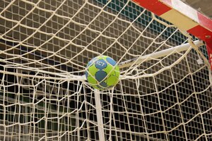 Ein blau-gr&uuml;ner Handball fliegt oben rechts in ein Tor. Das wei&szlig;e Tornetz f&auml;ngt den Ball ab.