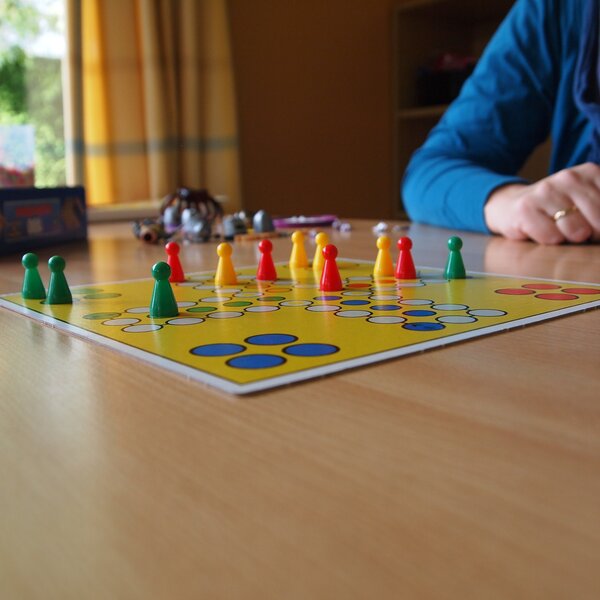 Bild vergr&ouml;&szlig;ern: Ein buntes Brettspiel liegt auf einem Holztisch. Auf dem gelben Spielbrett stehen rote, gelbe und gr&uuml;ne Spielfiguren. Eine Person in einem blauen Oberteil sitzt am Tisch.