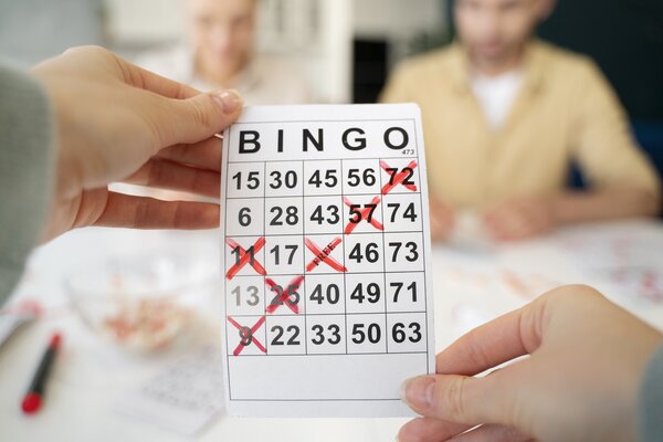 Bild vergr&ouml;&szlig;ern: Zwei H&auml;nde halten eine wei&szlig;e Bingo-Karte nah vor die Kamera. Einige Zahlen darauf sind mit roten Kreuzen markiert. Im unscharfen Hintergrund sitzen zwei Menschen an einem Tisch.