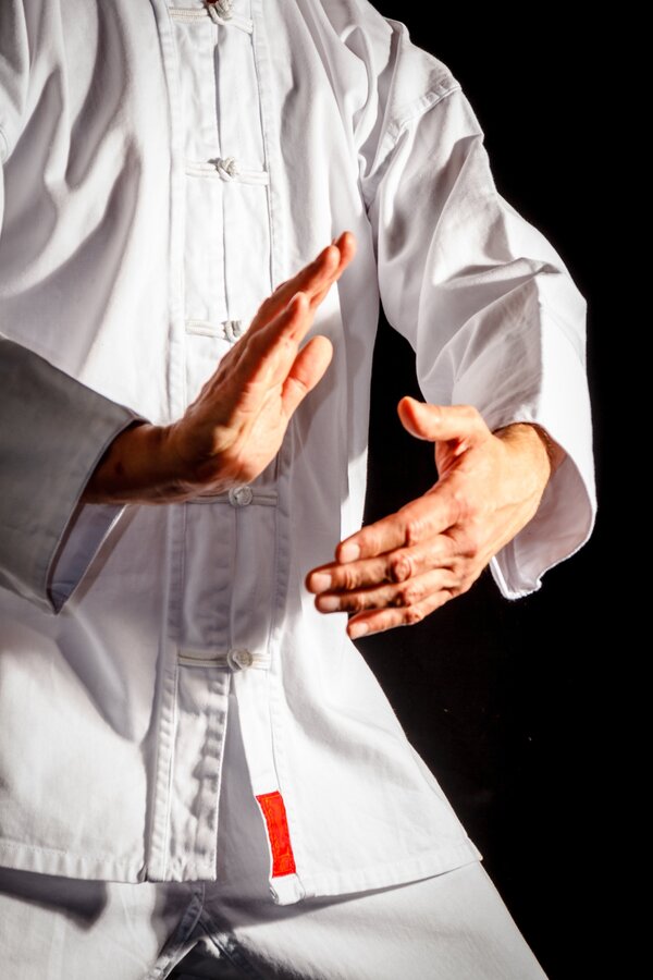Bild vergr&ouml;&szlig;ern: Eine Person in einem wei&szlig;en Kampfsportanzug macht eine ruhige Handbewegung im Stil von Tai-Chi. Zu sehen ist der Oberk&ouml;rper vor schwarzem Hintergrund.