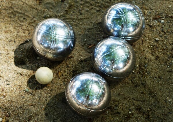 Bild vergr&ouml;&szlig;ern: Vier gro&szlig;e, silberne Metallkugeln und eine kleine helle Holzkugel liegen im Sand. Sie geh&ouml;ren zu einem Boule-Spiel. Die silbernen Kugeln gl&auml;nzen in der Sonne.