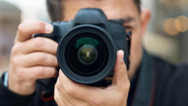 Bild vergr&ouml;&szlig;ern: Ein Mann h&auml;lt eine schwarze Kamera direkt vor sein Gesicht und fotografiert. Das gro&szlig;e Kamera-Objektiv zeigt nach vorne zum Betrachter.