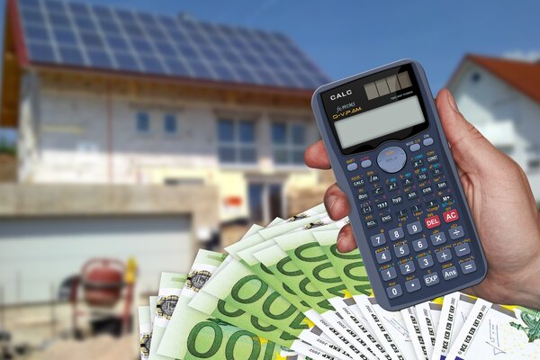 Eine Hand h&auml;lt einen Taschenrechner &uuml;ber vielen 100-Euro-Scheinen. Im unscharfen Hintergrund steht ein Haus mit einer Solaranlage auf dem Dach.