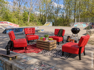Vier rote Sessel stehen im Kreis auf einem Skatepark. In der Mitte steht ein Tisch aus Holz mit Gl&auml;sern und einer Pflanze. Rechts steht ein schwarzer Kugelgrill. Im Hintergrund stehen B&auml;ume und Skaterampen mit Graffiti.