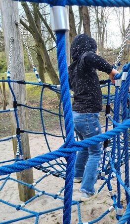 Bild vergr&ouml;&szlig;ern: Ein Kind klettert auf einem Spielplatz durch ein Netz aus dicken, blauen Seilen. Es tr&auml;gt einen dunklen Pullover mit kleinen Sternen und eine Jeans.