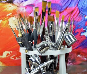 Viele Pinsel in verschiedenen Gr&ouml;&szlig;en und gl&auml;nzende Airbrush-Pistolen stecken in einem wei&szlig;en Halter. Der Hintergrund ist bunt mit leuchtenden Farben wie Pink, Gelb und Blau bemalt.