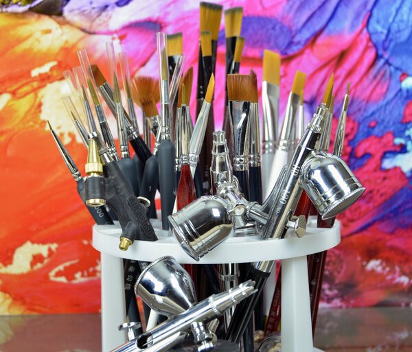 Bild vergr&ouml;&szlig;ern: Viele Pinsel in verschiedenen Gr&ouml;&szlig;en und gl&auml;nzende Airbrush-Pistolen stecken in einem wei&szlig;en Halter. Der Hintergrund ist bunt mit leuchtenden Farben wie Pink, Gelb und Blau bemalt.