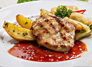 Ein gebratenes Fleischst&uuml;ck liegt auf einem wei&szlig;en Teller in roter So&szlig;e. Daneben liegen knusprige Kartoffelecken und kleine Tomatenscheiben.