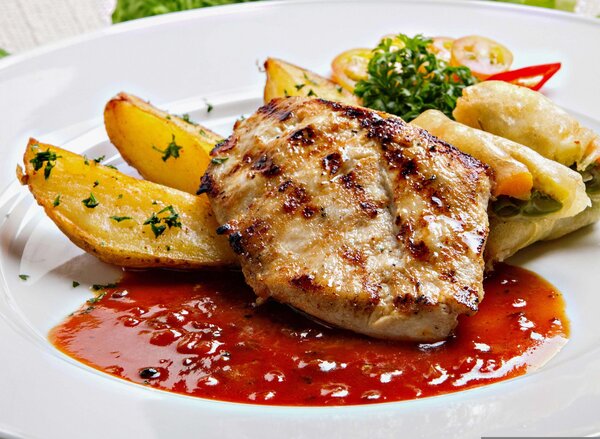 Bild vergr&ouml;&szlig;ern: Ein gebratenes Fleischst&uuml;ck liegt auf einem wei&szlig;en Teller in roter So&szlig;e. Daneben liegen knusprige Kartoffelecken und kleine Tomatenscheiben.