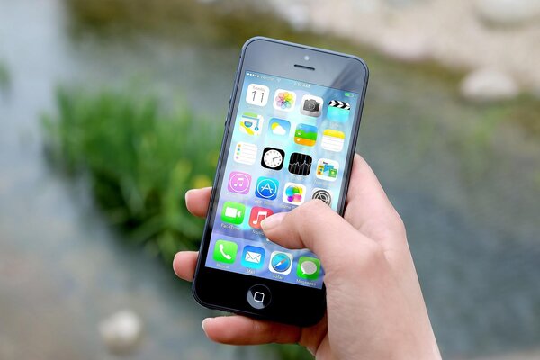 Bild vergr&ouml;&szlig;ern: Eine Hand h&auml;lt ein schwarzes Smartphone und tippt mit dem Daumen auf den bunten Bildschirm mit verschiedenen Apps. Der Hintergrund im Freien ist gr&uuml;n und unscharf.