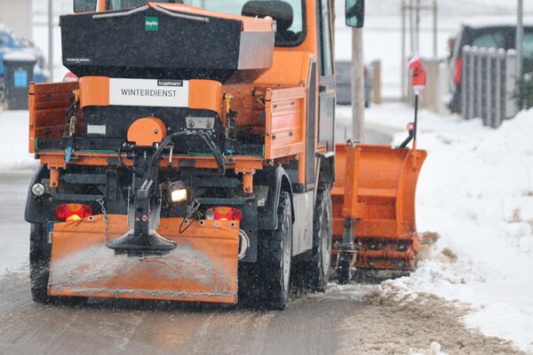 Ein oranges Winterdienst-Fahrzeug r&auml;umt Schnee von einer Stra&szlig;e. Vorne befindet sich ein Schneepflug, hinten ein Streuger&auml;t f&uuml;r Salz. Auf dem Fahrzeug steht in schwarzen Buchstaben das Wort Winterdienst.