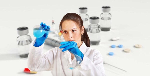 Eine Wissenschaftlerin im wei&szlig;en Kittel und mit blauen Handschuhen gie&szlig;t vorsichtig eine blaue Fl&uuml;ssigkeit in ein Glasgef&auml;&szlig;. Im Hintergrund sind Laborflaschen zu sehen.