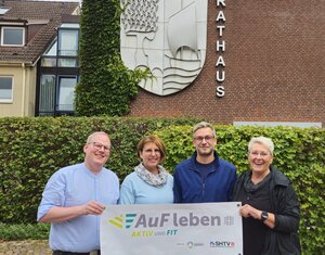 Vier l&auml;chelnde Personen stehen vor einem Rathaus aus Backstein. Zusammen halten sie ein Banner mit der Aufschrift AuF leben - AKTIV UND FIT.
