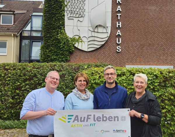 Vier l&auml;chelnde Personen stehen vor einem Rathaus aus Backstein. Zusammen halten sie ein Banner mit der Aufschrift AuF leben - AKTIV UND FIT.