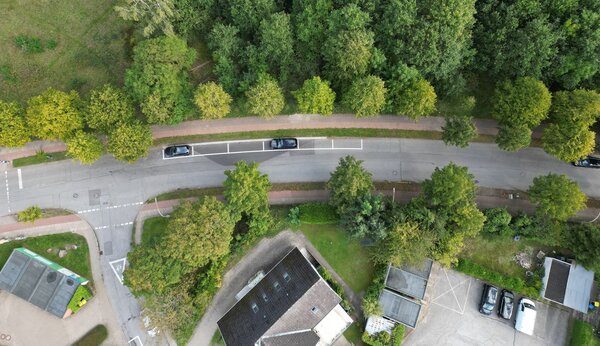 Eine Luftaufnahme von oben: Eine Stra&szlig;e f&uuml;hrt an vielen gr&uuml;nen B&auml;umen vorbei. Am Stra&szlig;enrand parken zwei dunkle Autos in markierten Parkbuchten. Unten im Bild ist das Dach eines Hauses zu sehen.