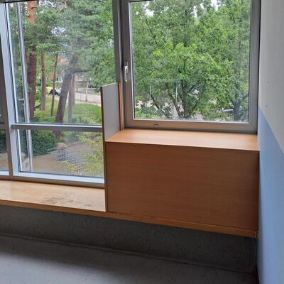 Ein gro&szlig;es Fenster gibt den Blick frei auf gr&uuml;ne B&auml;ume im Au&szlig;enbereich. Unter dem Fenster befindet sich eine niedrige, helle Holzbank.