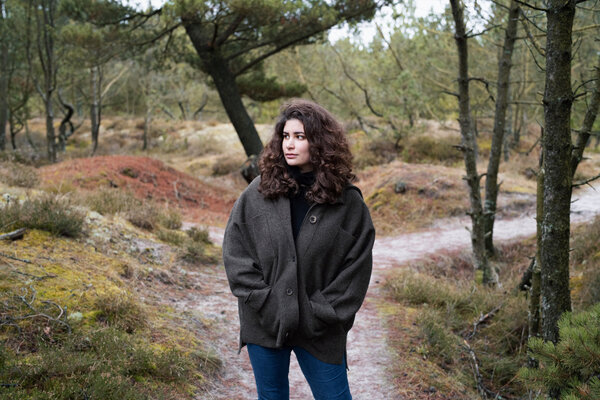 Bild vergr&ouml;&szlig;ern: Eine Frau mit dunklem, lockigem Haar und dunkler Jacke steht auf einem Waldweg.