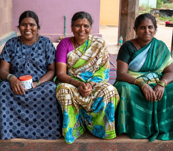 Bild vergr&ouml;&szlig;ern: Drei l&auml;chelnde indische Frauen sitzen nebeneinander vor einer lila Wand. Sie tragen farbenfrohe Gew&auml;nder. Die Frau links tr&auml;gt ein blaues gepunktetes Kleid, die anderen beiden Frauen tragen bunte Saris.