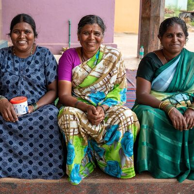 Drei l&auml;chelnde indische Frauen sitzen nebeneinander vor einer lila Wand. Sie tragen farbenfrohe Gew&auml;nder. Die Frau links tr&auml;gt ein blaues gepunktetes Kleid, die anderen beiden Frauen tragen bunte Saris.