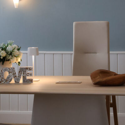 Bild vergr&ouml;&szlig;ern: Ein heller Tisch steht vor einer blau-wei&szlig;en Wand. Auf dem Tisch stehen wei&szlig;e Blumen, eine kleine Lampe, eine Holzschale und die Buchstaben LOVE. Hinter dem Tisch steht ein hoher, heller Stuhl.